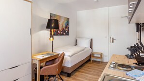 Habitación de hotel compacta con cama, escritorio y silla, una mesita, un cuadro en la pared y una kitchenette con cafetera.