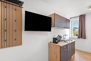 A modern kitchen with a flat-screen TV mounted on the wall, wooden cabinets, a countertop with a coffee machine, and a window with a view of the outdoors. - Studio Apartment 'Möbliertes Radebeul' with Balcony and Wi-Fi (Radebeul)