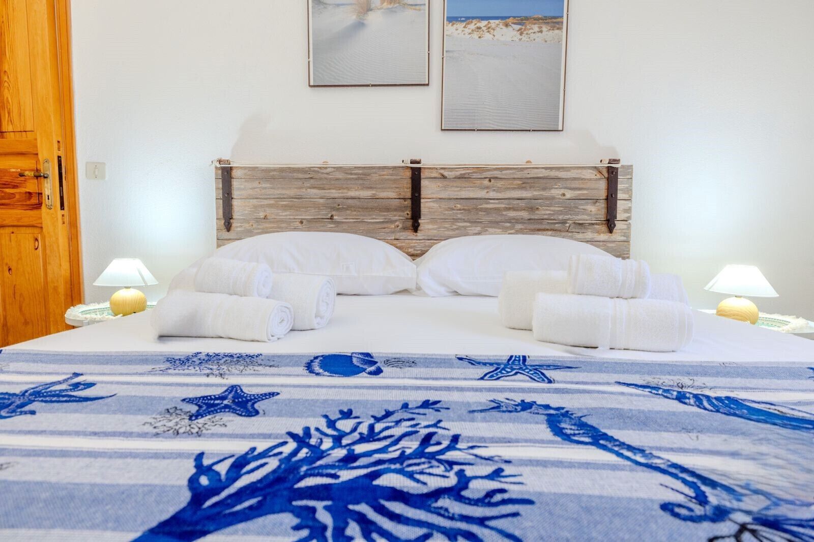Quarto de hotel com cabeceira de madeira, roupa de cama branca e colcha com padrão azul.