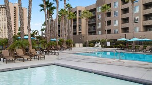 Ein Hotelpoolbereich mit Liegestühlen und Sonnenschirmen, umgeben von hohen Palmen und einem mehrstöckigen Gebäude mit Balkonen.