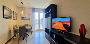 Hotellihuone, jossa on ruokailutila, seinälle asennettu televisio ja punainen maljakko pöydällä.