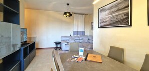 A modern living room with a grey sofa, a dining table with a magazine and a book, a TV on a stand, and a large framed picture on the wall. - City / Village Apartment in Viareggio with 2 bedrooms sleeps 4 (Viareggio)