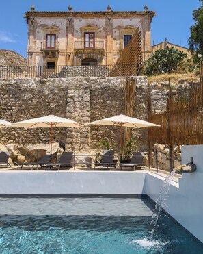 A pool area with a stone wall, two umbrellas, and lounge chairs. - Baglio Capo  (siculiana)