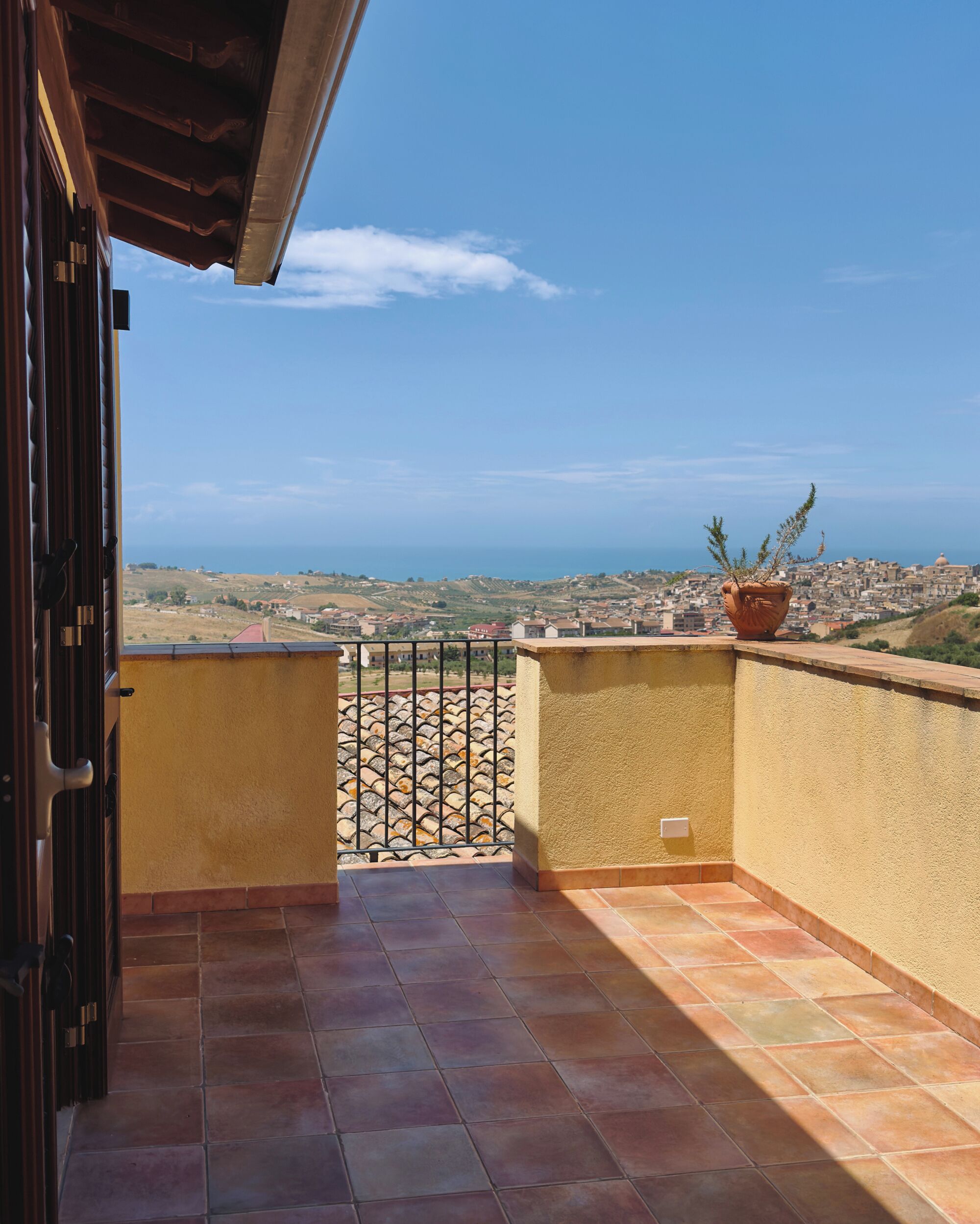 A balcony with a view of a coastal town, a potted plant, and a tiled floor.