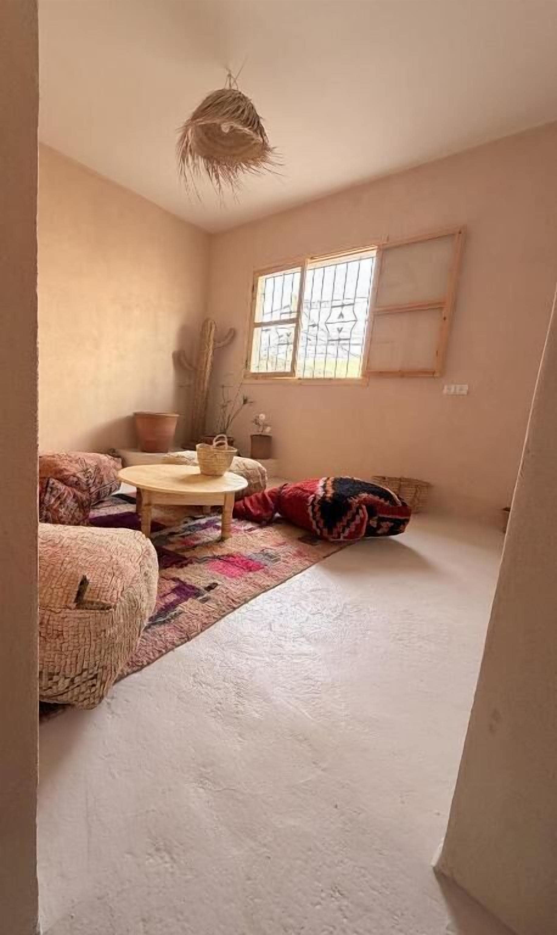 A room with a low ceiling, a window with bars, a cactus, a woven pouf, a round wooden table, and a patterned rug.