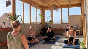 A group of people sitting on yoga mats in a room with a wooden ceiling and large windows. - Sines Surfmaroc (Aourir)