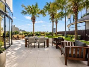 Eine Außenpatio-Terrasse mit Tischen und Stühlen, Palmen und einem modernen Gebäude im Hintergrund.