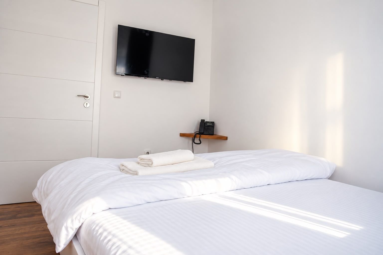Habitación de hotel con cama, televisor de pantalla plana montado en la pared, teléfono sobre un estante de madera y puerta con manija.
