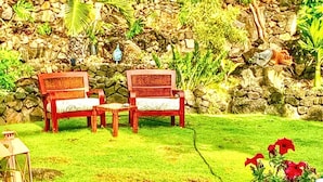 A garden with a stone wall, two wooden benches, a wicker chair, and a potted plant with red flowers. - Hale Akala - Huahine (Shared Bathroom) (Kailua-Kona)