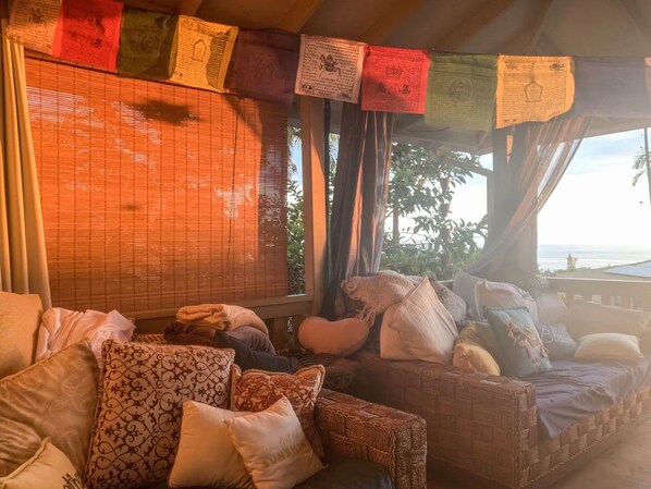 A cozy seating area with a wicker sofa and cushions, a window with blinds, and a view of the outdoors.
