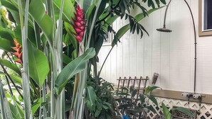 A patio area with a wooden bench, a metal chair, and a variety of tropical plants including a banana tree and a red flower. - Hale Akala - Surf Ohana (Kailua-Kona)