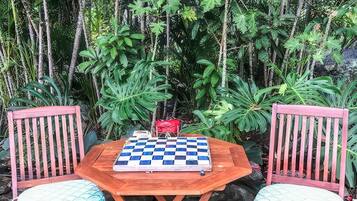 A checkered game board on a wooden table with two chairs, set outdoors with dense foliage in the background.