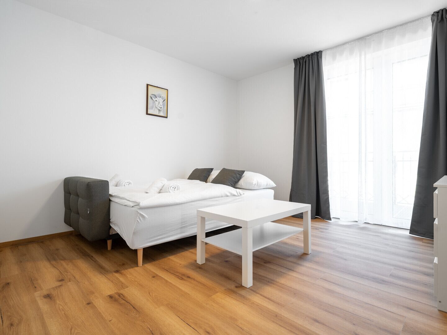Ein schlicht eingerichteter Raum mit einem Bett, einem kleinen Tisch, einer Couch und einem einzelnen Bild in einem Rahmen an der Wand.