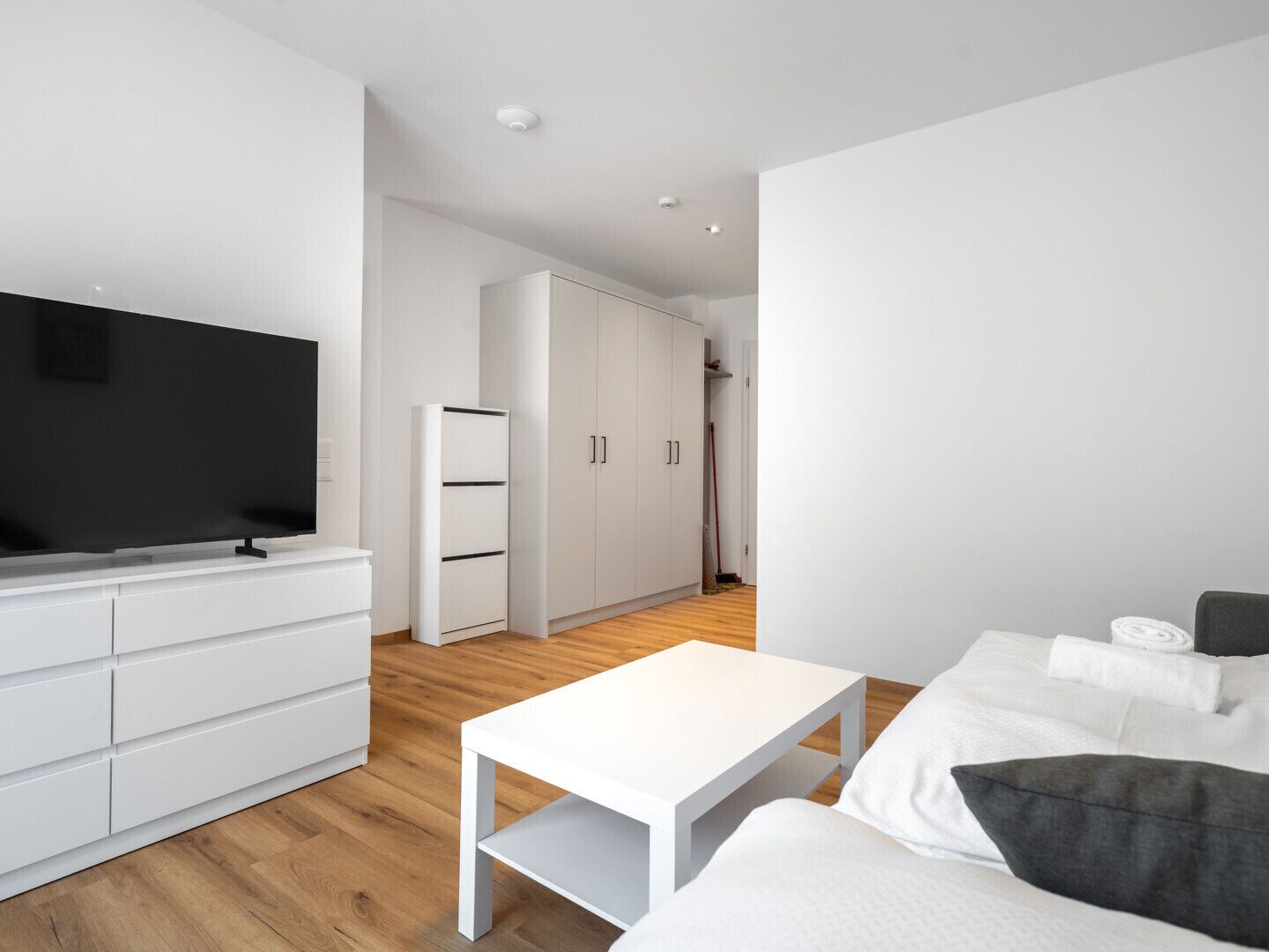 A modern living room with a flat-screen TV, white furniture, a wooden floor, and a large wardrobe.