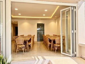A dining area with a long wooden table and wicker chairs, a glass door leading to an outdoor space, and recessed ceiling lighting.