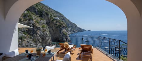 A terrace with a table set for dining, chairs, and a view of the sea.