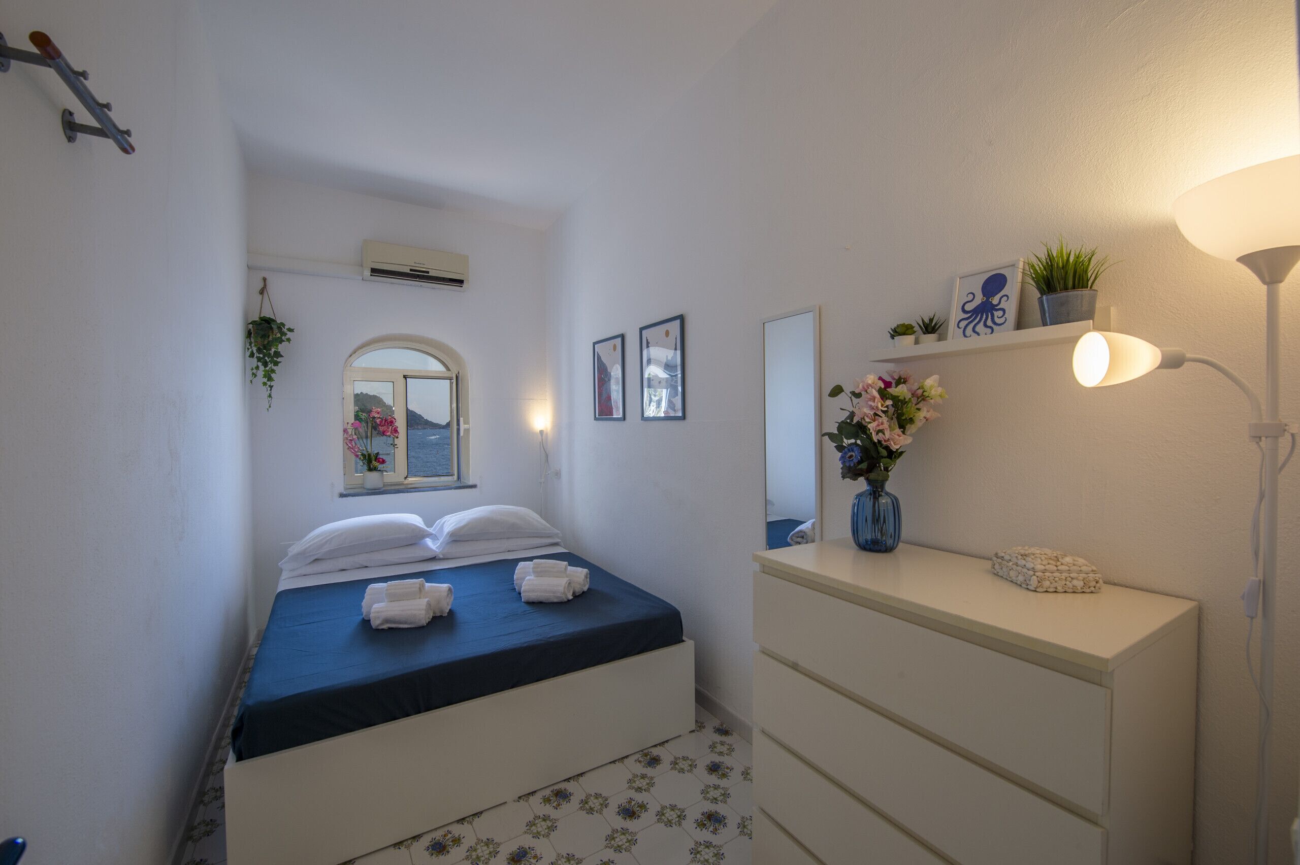Une chambre d’hôtel comprenant un lit, une table de chevet, un miroir, un climatiseur fixé au mur et une fenêtre donnant sur la mer.