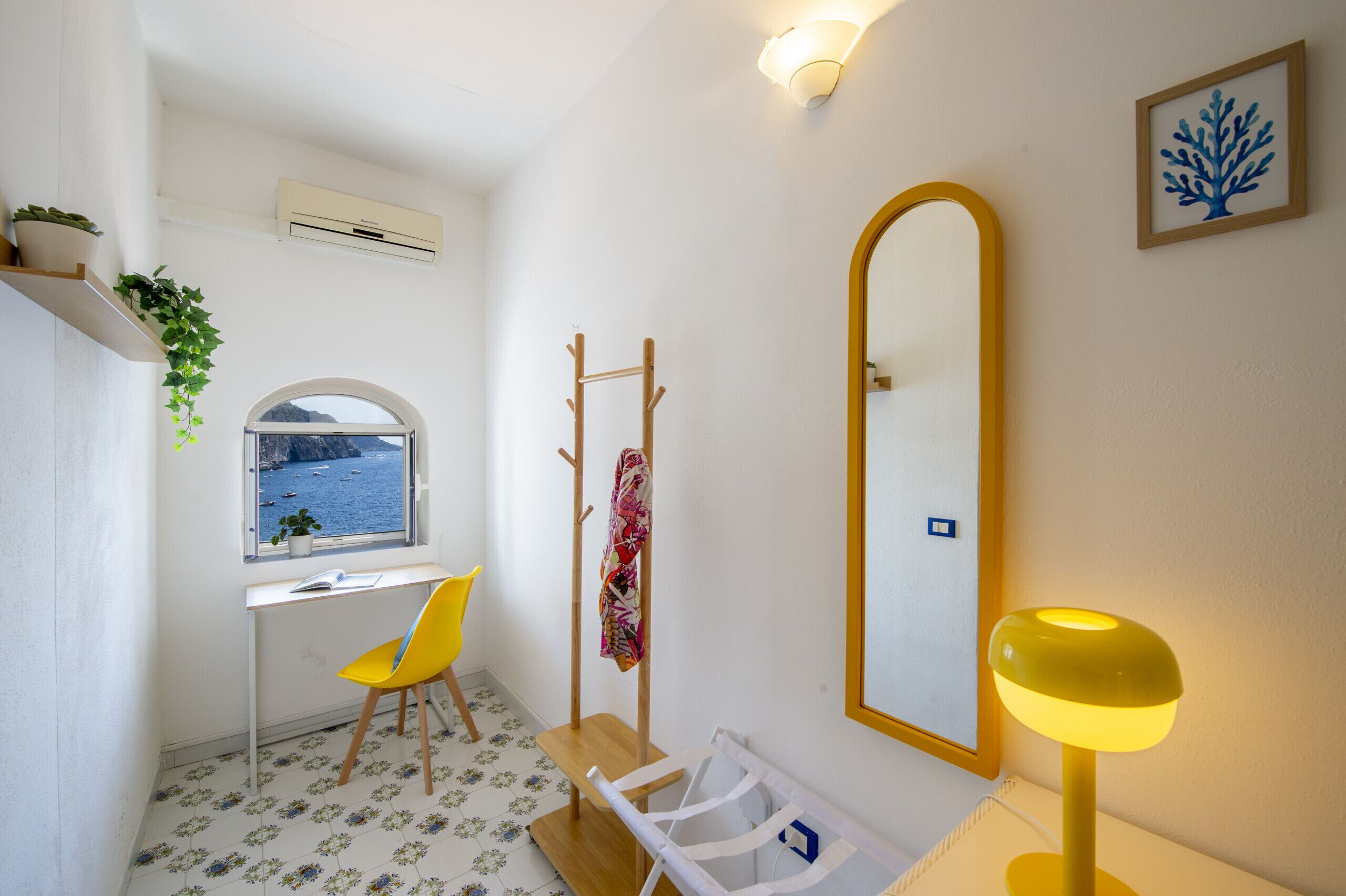 Une petite salle de bain avec une fenêtre, un miroir, une chaise jaune, une petite table et une plante.