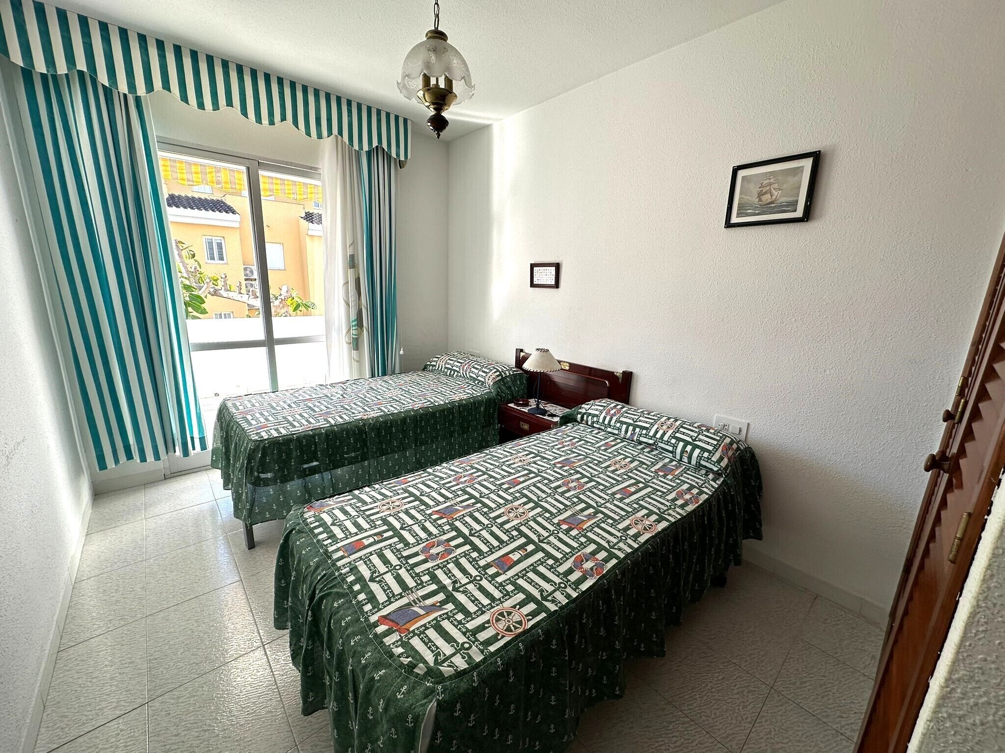 Una habitación con dos camas, una ventana con cortinas, un cuadro en la pared y una puerta.