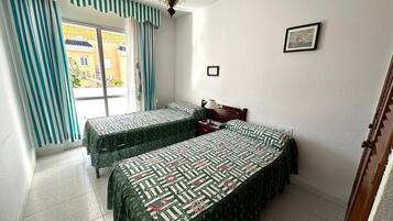 Una habitación con dos camas, una ventana con cortinas, un cuadro en la pared y una puerta.