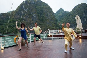 A group of people performing on a wooden deck with a view of green mountains in the background. - Orchid Luxury Cruise (Bai Chay)