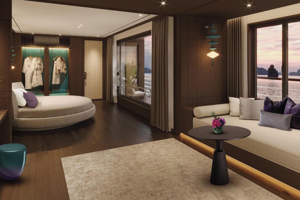 A modern hotel room with a curved sofa, a round table, a wardrobe with hanging clothes, and a view of the sea.