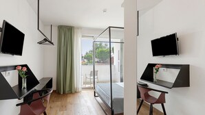 Een moderne kamer met een bed, twee bureaus met computers, een televisie en een raam met gordijnen.