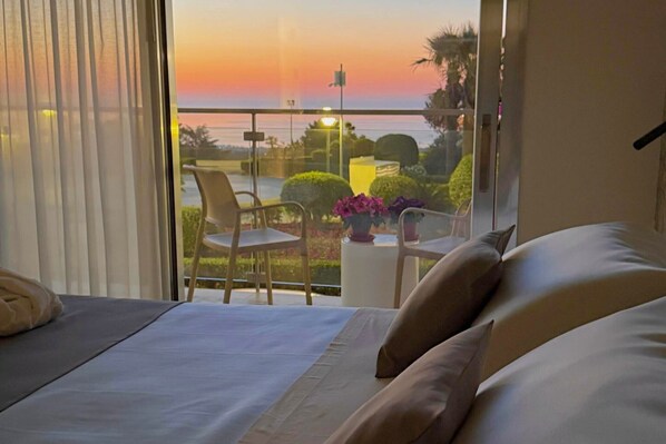 Ein Hotelzimmer mit Bett, Stuhl, kleinem Tisch mit Blumenvase und einem Balkon mit Sonnenuntergangsblick.