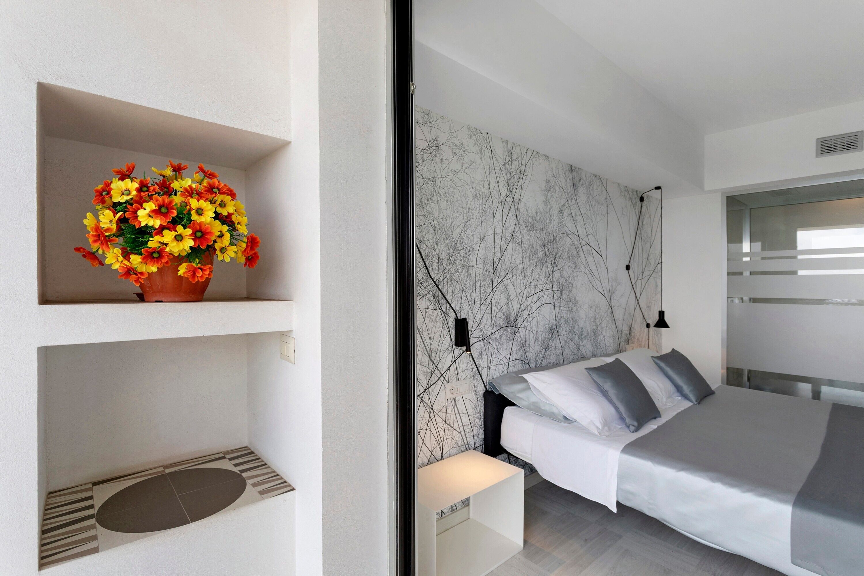 A modern bedroom with a bed, a marble-patterned headboard, a flower arrangement on a shelf, and a wall-mounted light fixture.