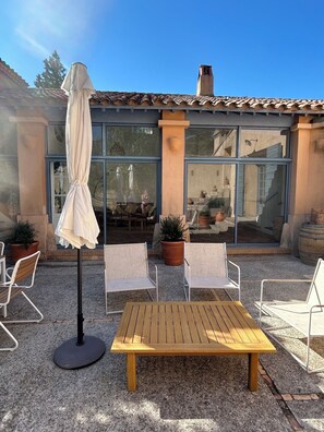 Um pátio externo com mesa de madeira e duas cadeiras, guarda-sol e plantas em vasos.