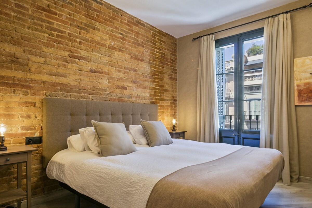 Ein Schlafzimmer mit Backsteinwand, einem großen Bett mit vielen Kissen, einem Nachttisch mit Lampe und einem Fenster mit Vorhängen.