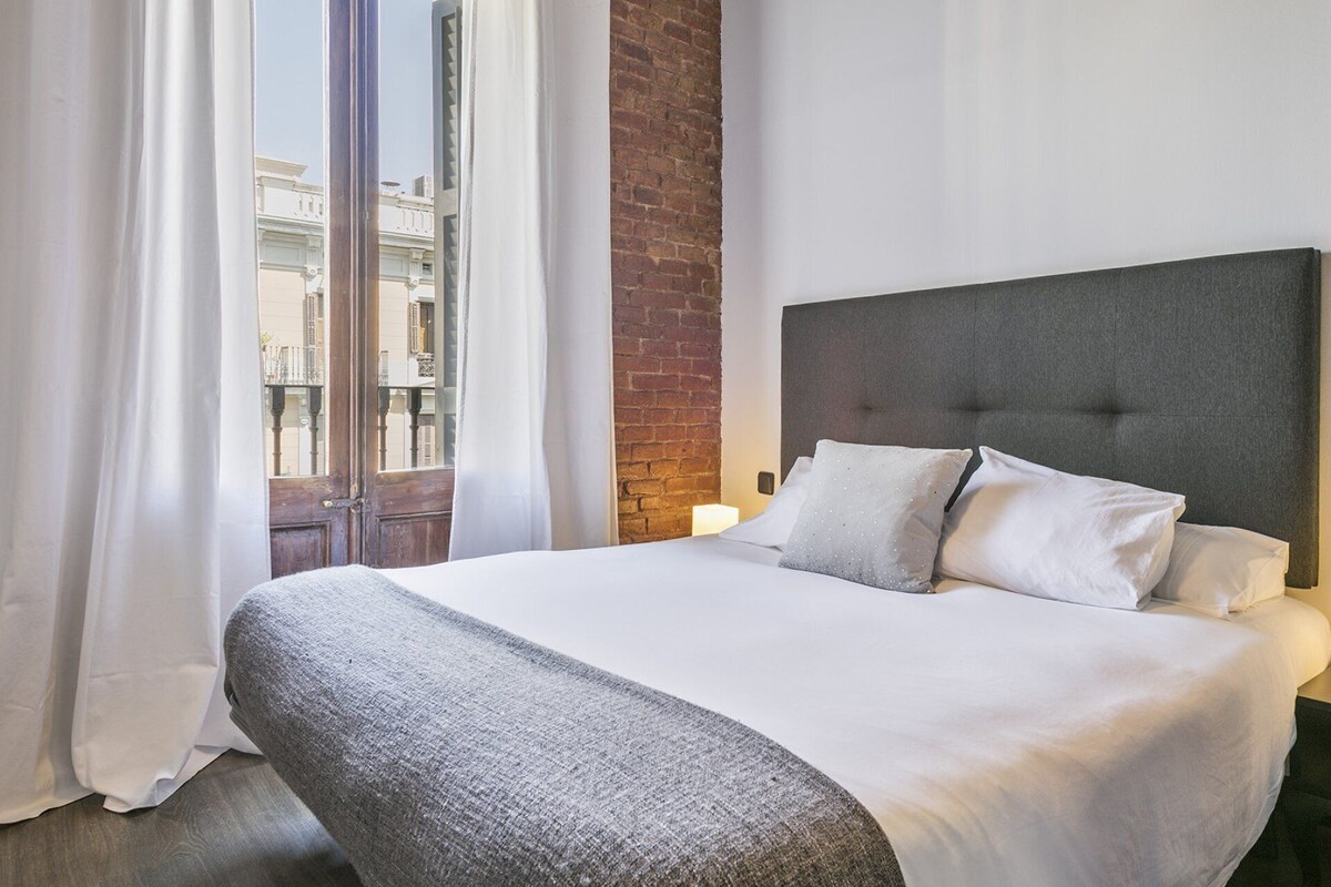 Ein Hotelzimmer mit einem großen Bett, weißen Vorhängen, einer Ziegelwand und Blick auf Gebäude.