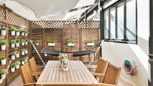Eine Terrasse im Freien mit einem Holztisch und -stühlen, umgeben von Blumentöpfen, einer Hängematte und Blick auf die Außenfassade des Gebäudes.