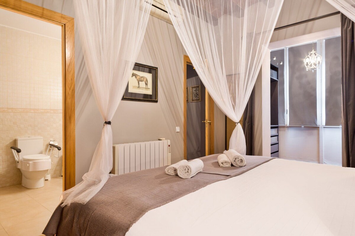 Ein Schlafzimmer mit einem großen Bett, einem Nachttisch, einem gerahmten Bild an der Wand und einem Fenster mit Vorhängen.