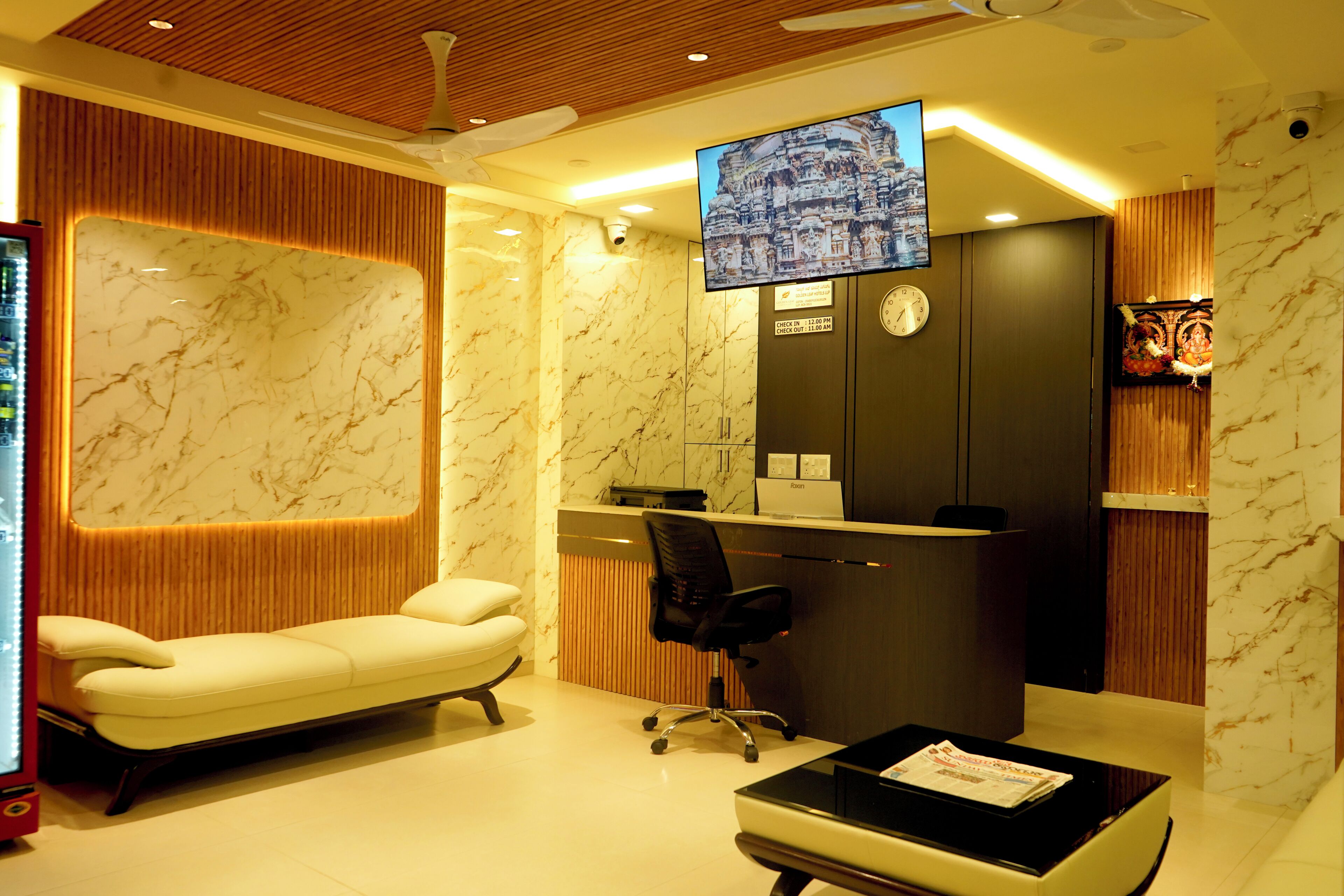Zona de recepción de hotel con mostrador, un sofá, una silla, un televisor montado en la pared y un reloj.