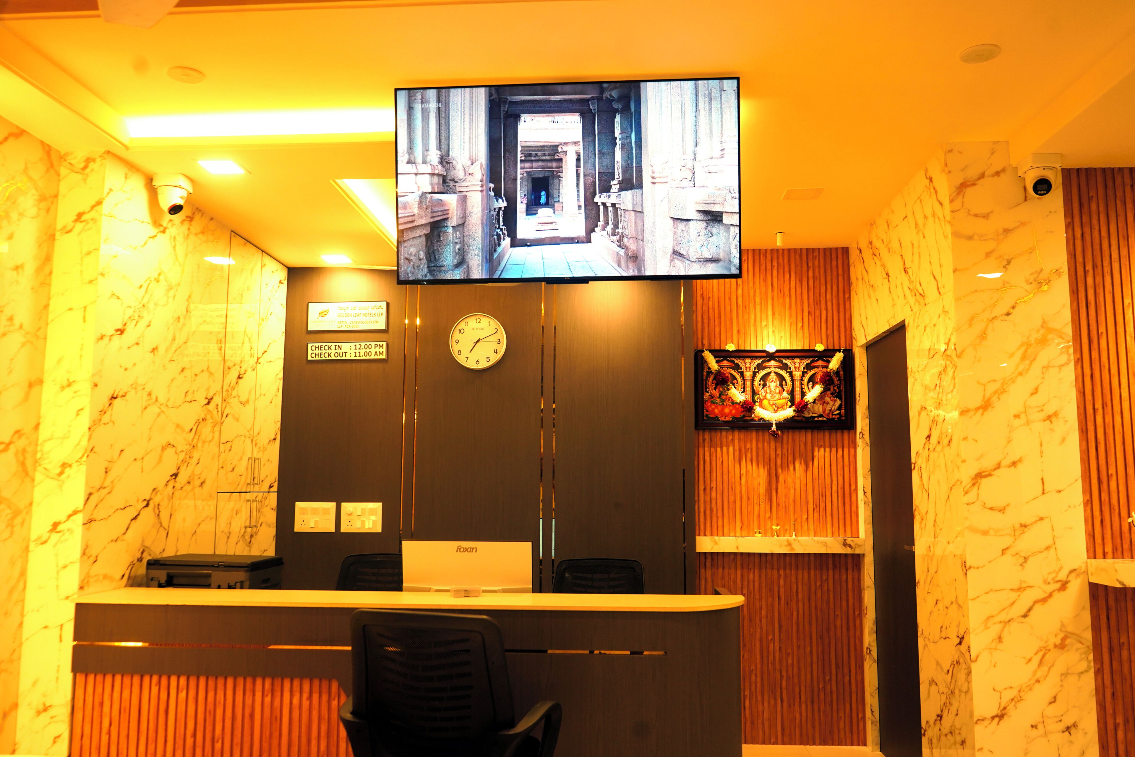 Ein Hotel-Empfangsbereich mit an der Wand montiertem Fernseher, der eine Szene zeigt, einer Uhr und einem Empfangstresen mit einem Stuhl davor.