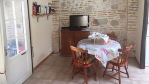 A dining area with a table set for two, a television on a wooden cabinet, and a stone wall. - Charming gite with a private pool in the French countryside (Saint-Étienne-de-Fougères)