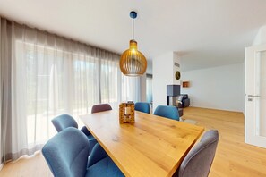 A modern living room with a wooden dining table, blue chairs, a hanging light fixture, and a large window with sheer curtains. - Chalet AlpspitzBlick – Alpine Chalet with Sauna & Garden, Garmisch-Partenkirchen (Garmisch-Partenkirchen)