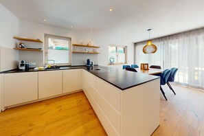 A modern kitchen with a central island, wooden flooring, and a dining area with blue chairs. - Chalet AlpspitzBlick – Alpine Chalet with Sauna & Garden, Garmisch-Partenkirchen (Garmisch-Partenkirchen)