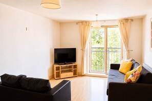 A living room with a flat-screen TV on a wooden stand, a sofa with cushions, and a balcony with curtains. - Modern Brindley Point Apt, Near Canals & Broad (West Midlands)