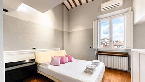 A bedroom with a bed, pillows, a radiator, and a window with a view of buildings. - Vigna Luxury Florence (Firenze)