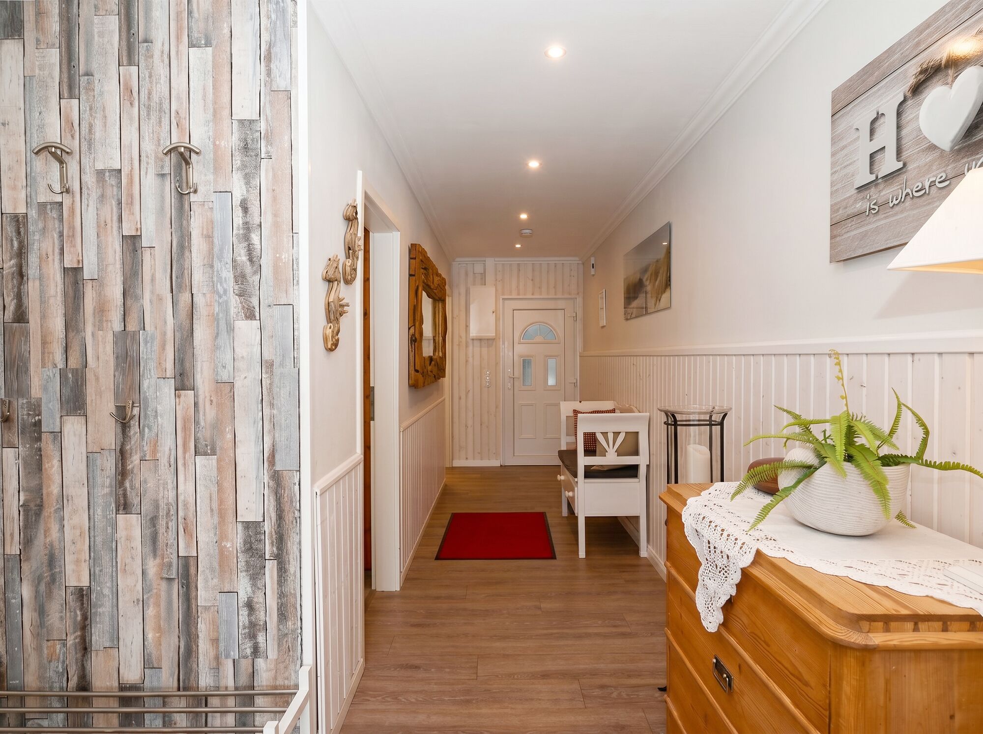 A hallway with a wooden floor, a red rug, a white chair, a wooden dresser with a plant, and a wall with a 'Home is where love is' sign.