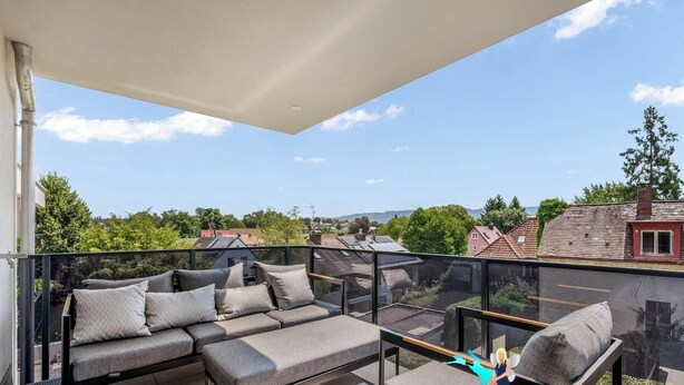 Un balcón con sofá y cojines, barandilla de vidrio y vista a una zona residencial.