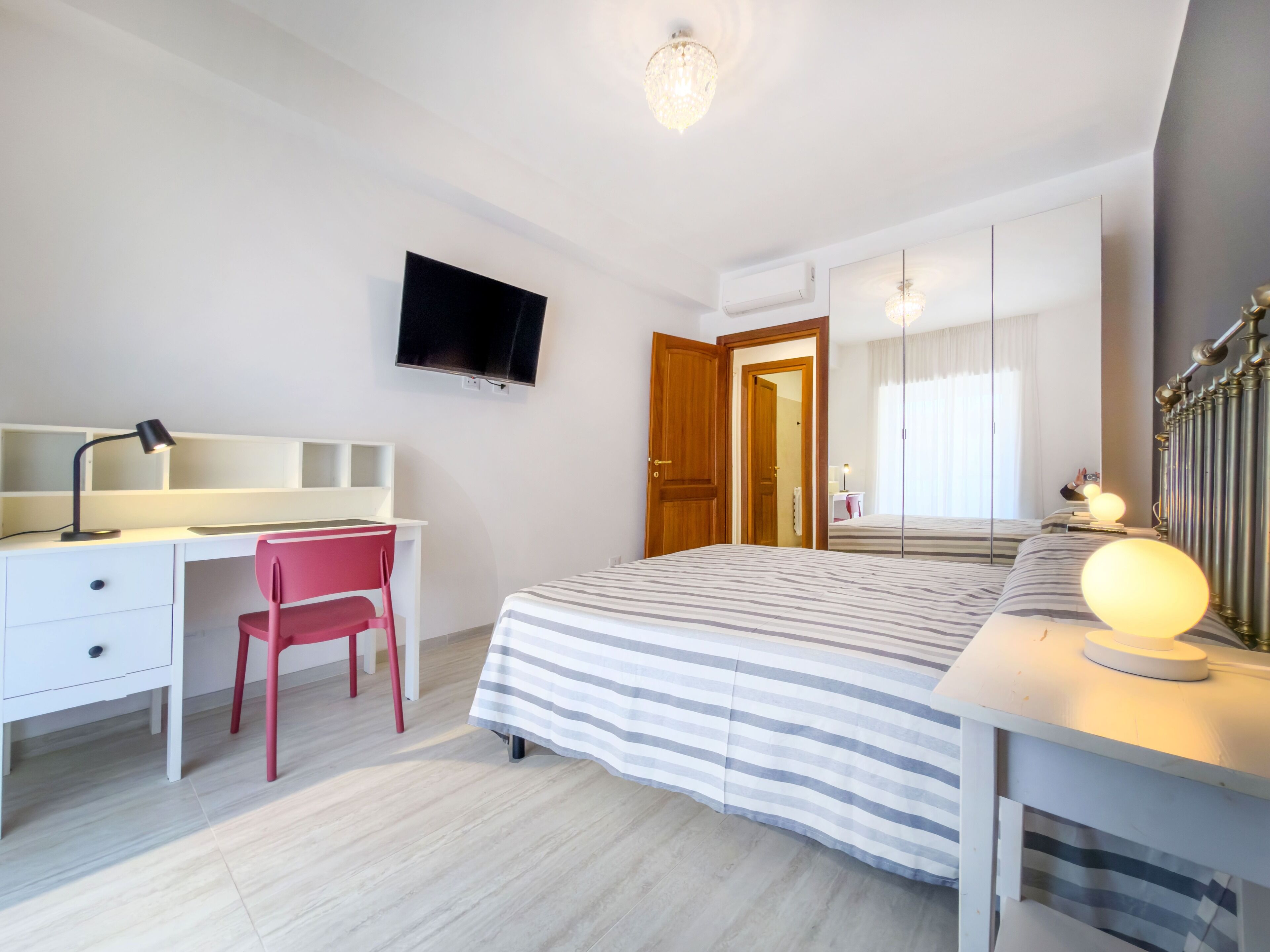 A bedroom with a bed, a desk with a chair, a television, a wardrobe, and a mirror.