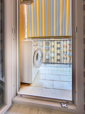 Ein Waschbereich mit Waschmaschine, einem Fenster mit Blick auf einen Balkon und einem gestreiften Markisenstoff.