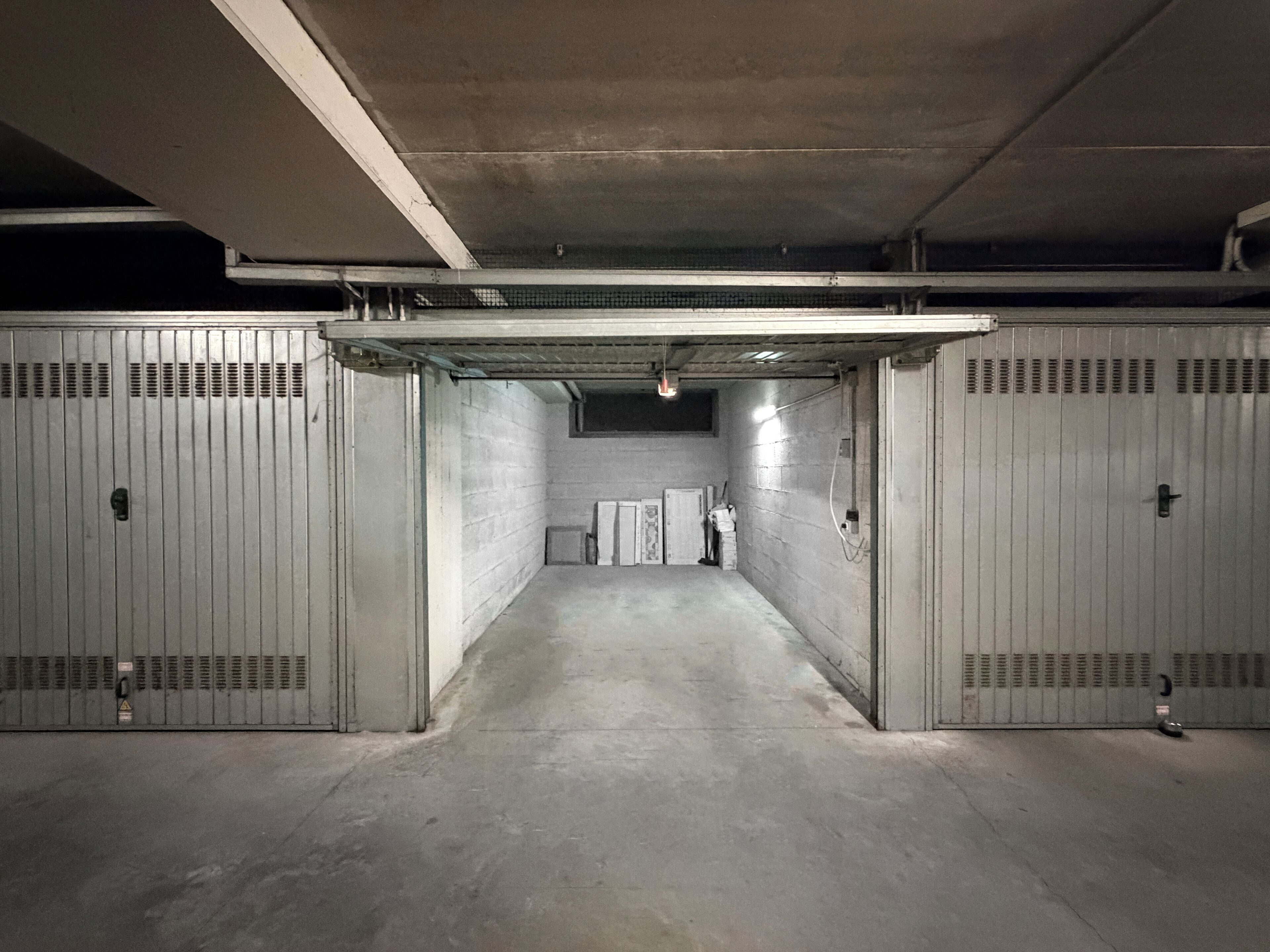 A parking garage with concrete walls and metal doors, featuring a central walkway and a row of storage units.