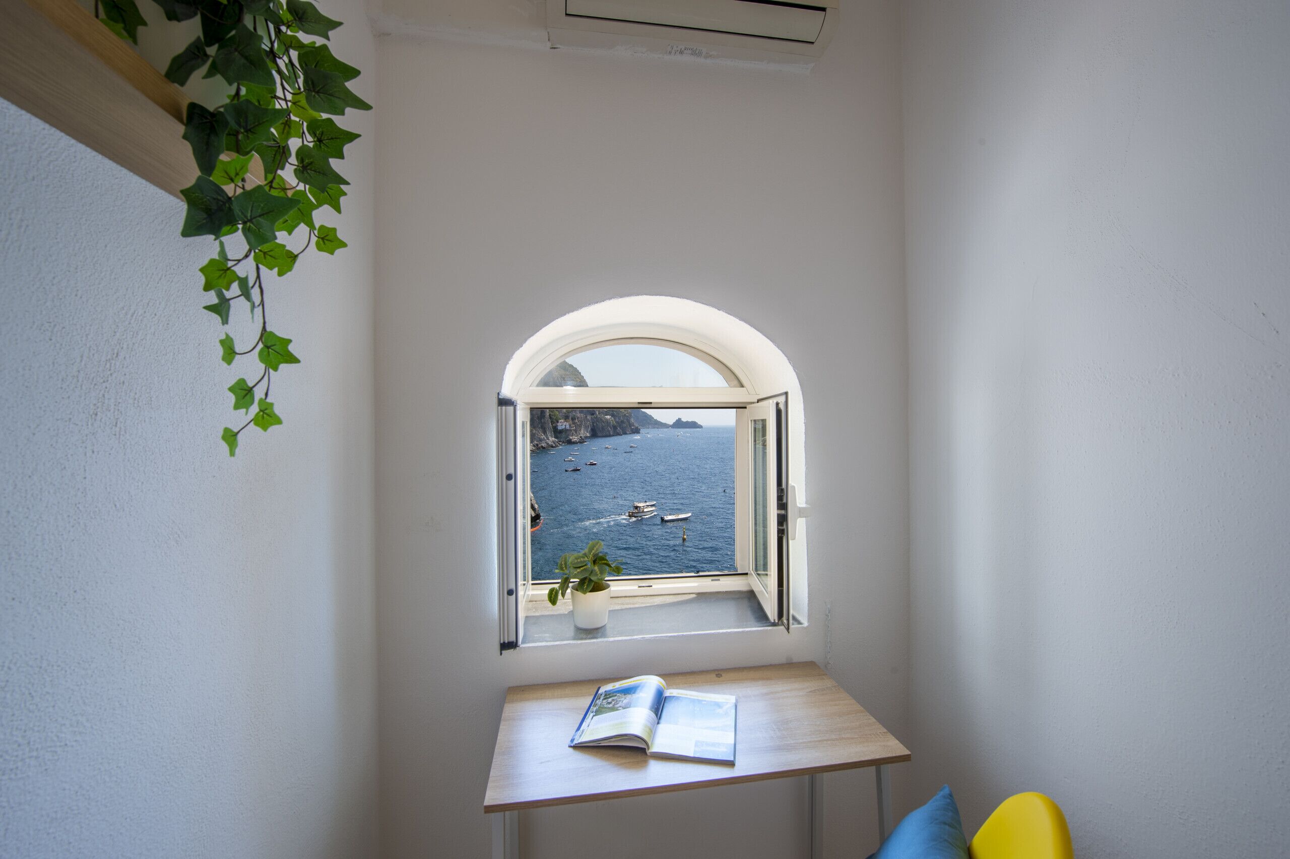 Une petite pièce avec une fenêtre donnant sur un plan d’eau et des bateaux, une table avec un livre et une plante en pot.