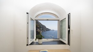 A room with a window overlooking a coastal landscape, a potted plant on the windowsill, and an open book on the table. - Casa Albachiara Waterfront Just Steps From the Beach (Praiano)