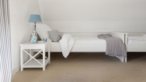 Ein helles Schlafzimmer im Loft-Stil mit schrägem Dach, einem kleinen Nachttisch mit Lampe, einem weißen Bett mit grauer Decke und einem gestreiften Kissen.