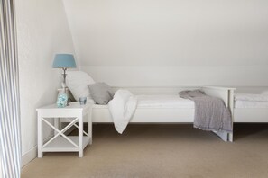 Ein helles Schlafzimmer im Loft-Stil mit schrägem Dach, einem kleinen Nachttisch mit Lampe, einem weißen Bett mit grauer Decke und einem gestreiften Kissen.